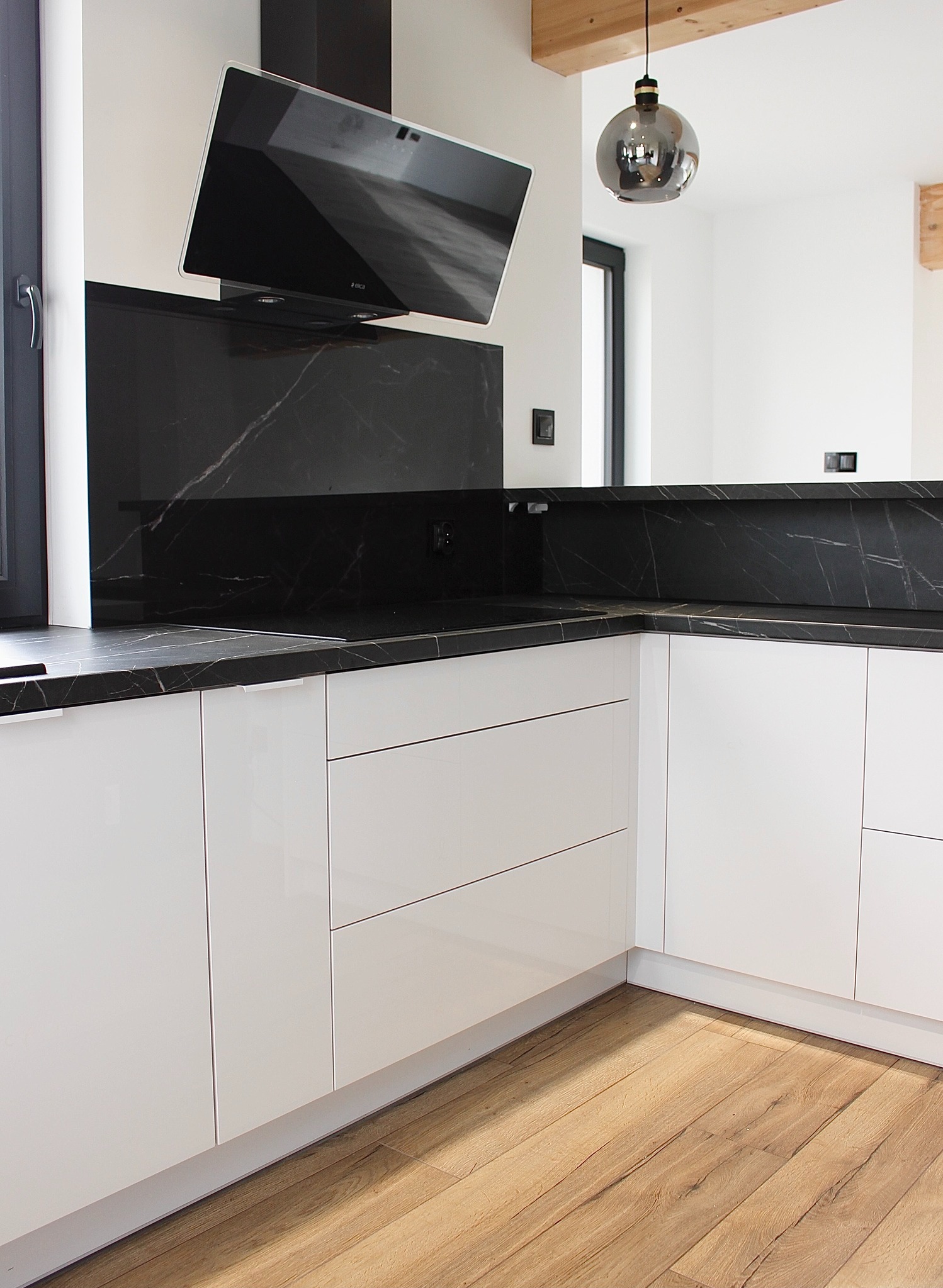 Nowoczesna kuchnia z białymi, lakierowanymi szafkami, czarnym blatem z marmurowym wzorem i drewnianą podłogą. Minimalistyczny okap i designerska lampa.