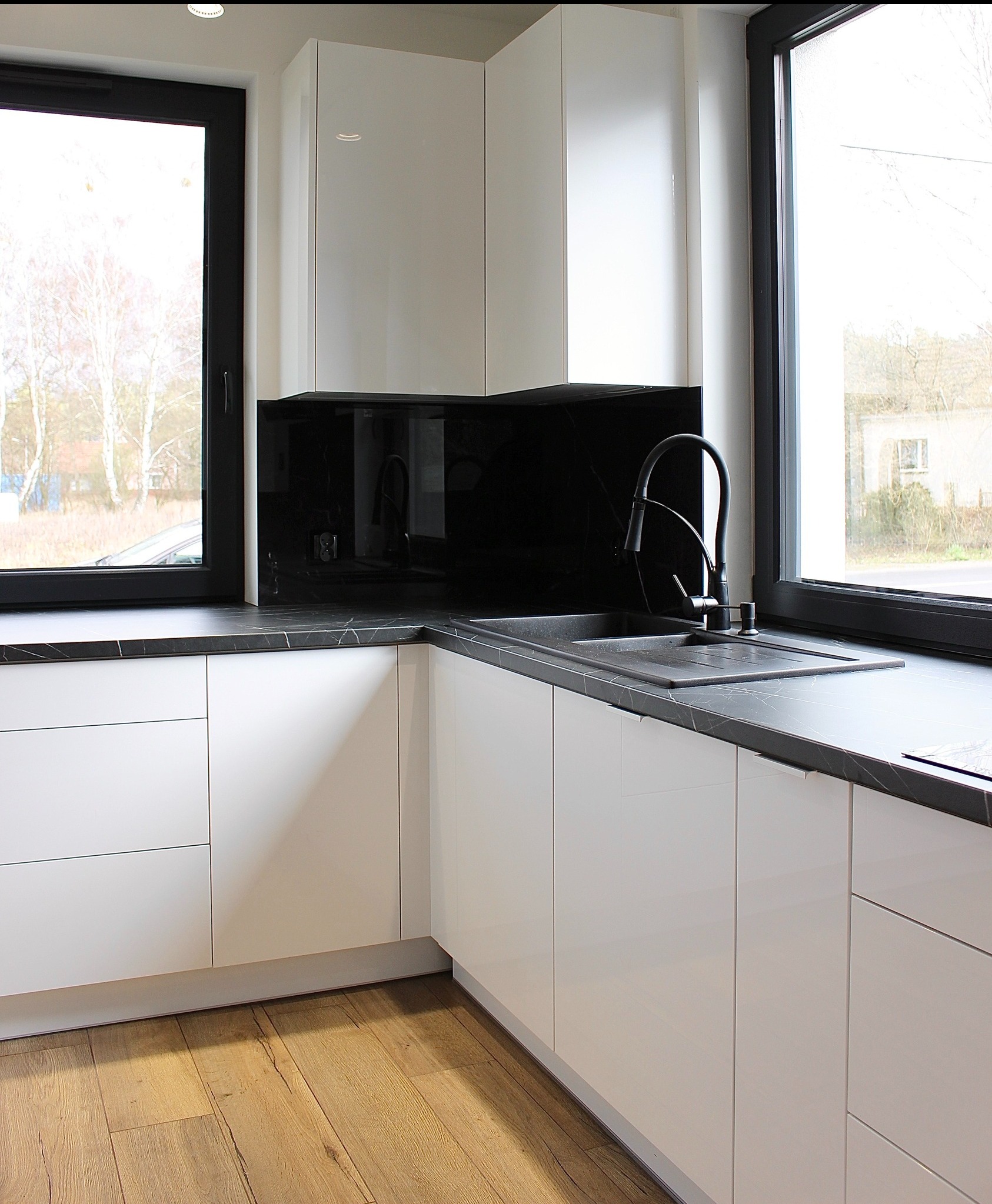 Nowoczesna kuchnia z białymi, lakierowanymi szafkami, czarnym blatem z marmurowym wzorem i drewnianą podłogą. Widok na okno z widokiem na zewnątrz.