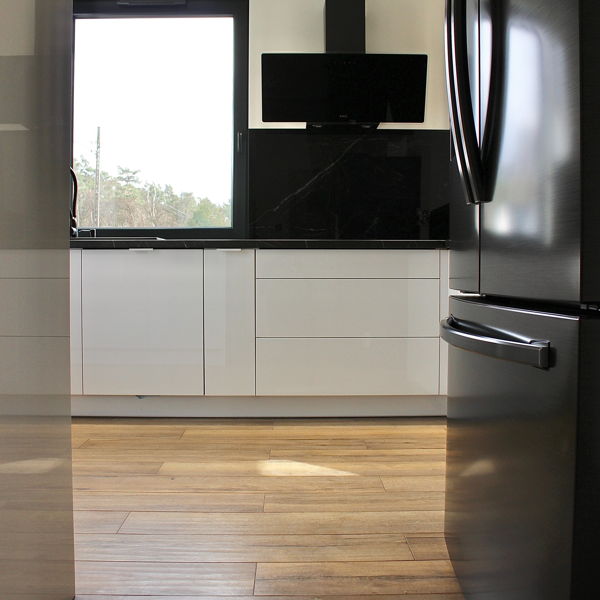 Nowoczesna kuchnia z białymi szafkami, czarnym blatem i okapem. Parkiet w odcieniach brązu. Częściowo widoczna lodówka ze stali nierdzewnej.