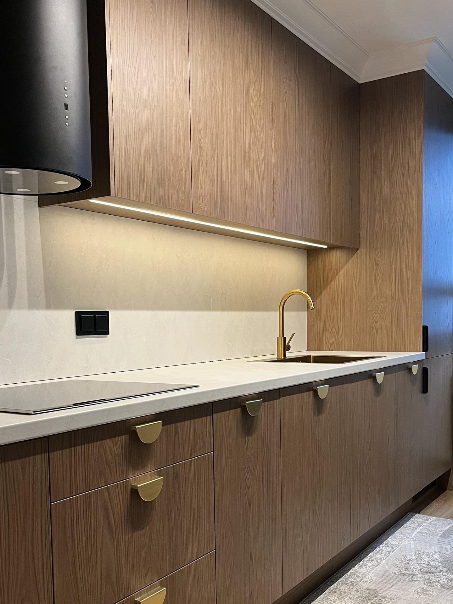 Nowoczesna kuchnia z drewnianymi frontami, złotą armaturą i oświetleniem LED pod szafkami. Minimalistyczny design z czarnym okapem i płytą indukcyjną.