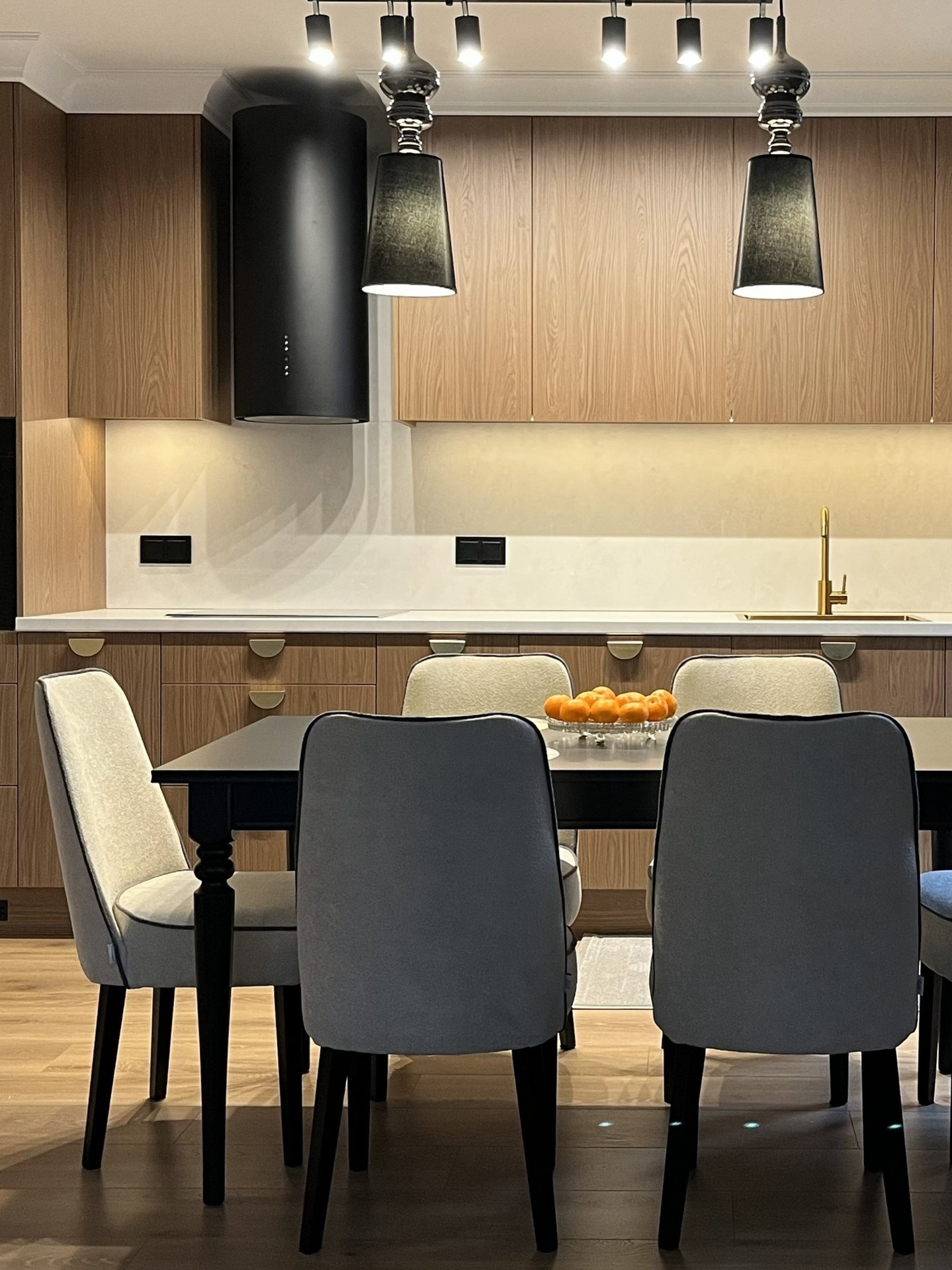 Nowoczesna kuchnia z drewnianymi szafkami, czarnym okapem i lampami. Na stole misa z owocami. Krzesła z szarym obiciem. Ciepłe oświetlenie.