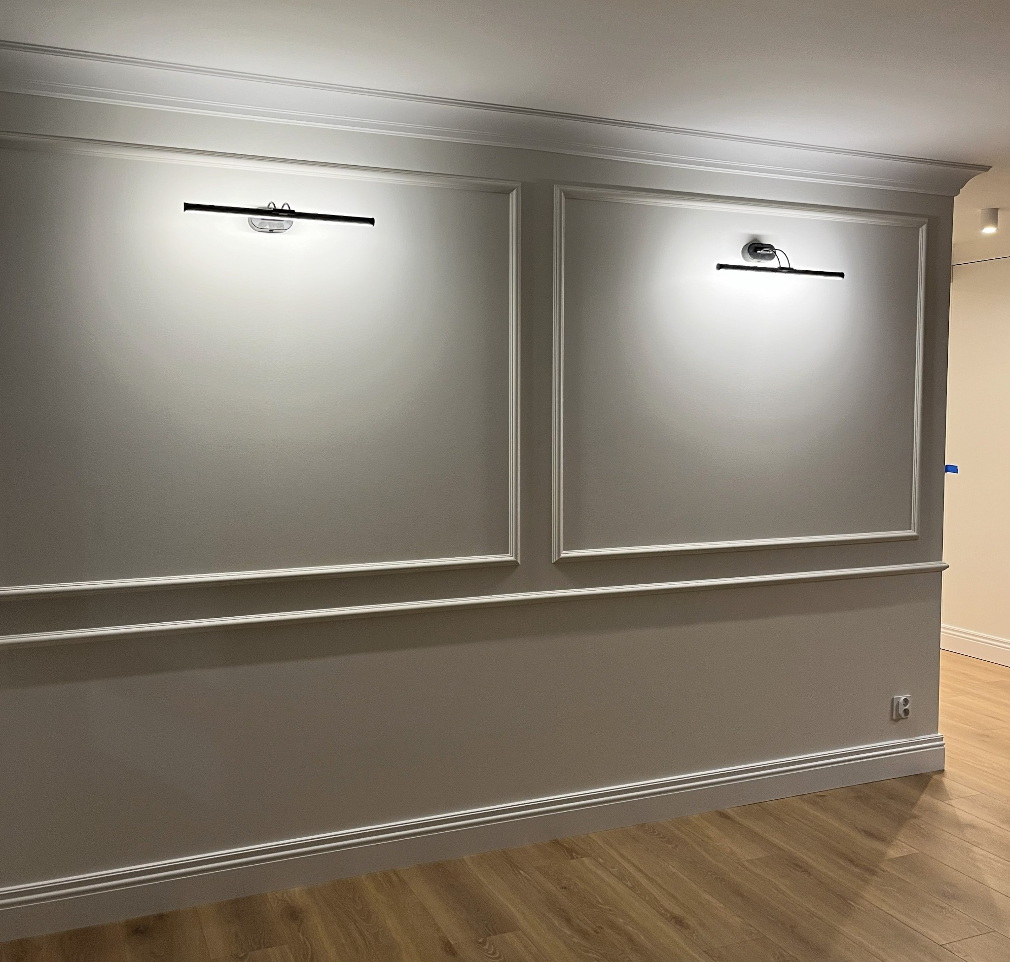 Szara ściana z dekoracyjnymi panelami i dwoma nowoczesnymi lampami ściennymi, oświetlającymi powierzchnię. Parkiet na podłodze, widoczne gniazdko elektryczne.