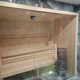 Nowoczesna, jasna kabina z drewnianym wnętrzem, szklanymi drzwiami i kamiennym piecem, otoczona marmurowymi płytkami.