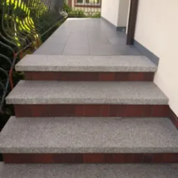 Zewnętrzne schody wejściowe wykończone szarą kostką granitową z czerwoną cegłą, prowadzące na balkon z szarą terakotą i czarną, metalową barierką.