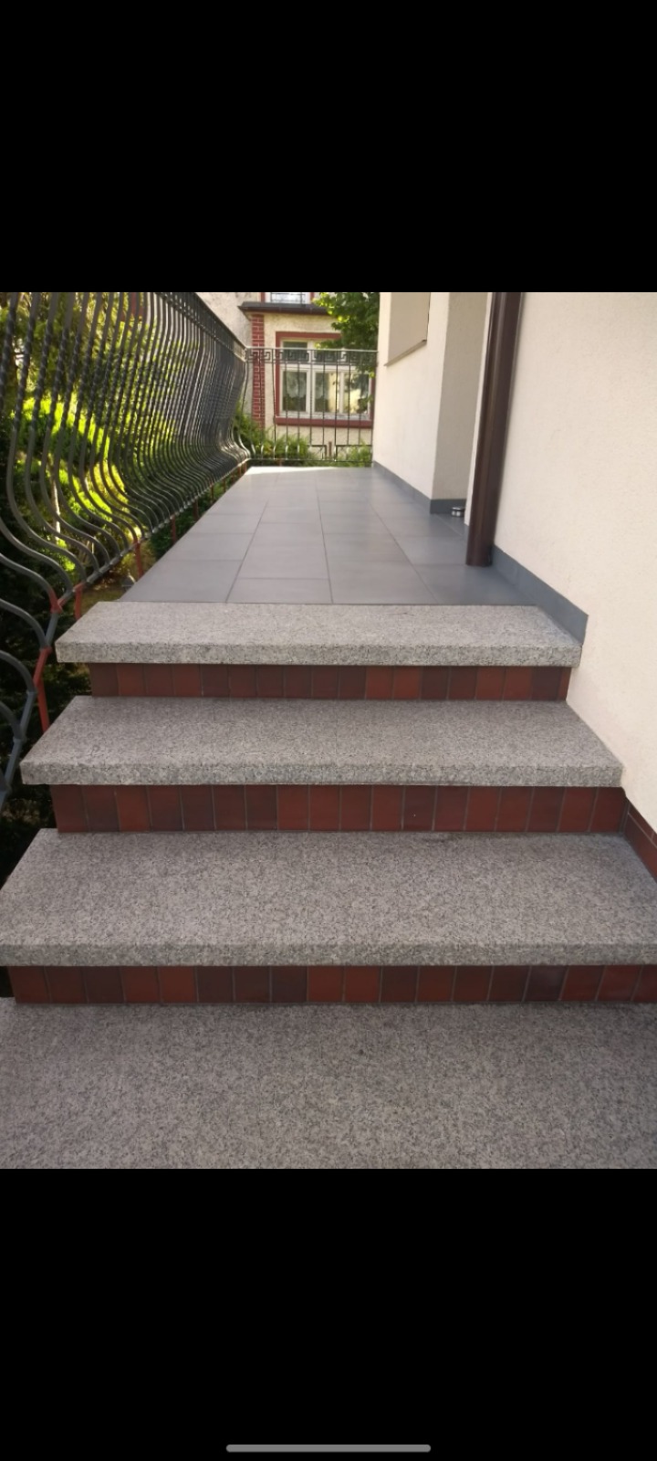 Zewnętrzne schody wejściowe wykończone szarą kostką granitową z czerwoną cegłą, prowadzące na balkon z szarą terakotą i czarną, metalową barierką.