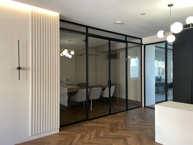 Nowoczesne biuro z przeszklonymi ścianami działowymi w czarnych ramach, podłoga z parkietu w jodełkę, ściana z dekoracyjnymi panelami i minimalistyczny zegar.