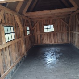 Damian Szlaga - Surowe wnętrze drewnianej konstrukcji z dwoma małymi oknami i posadzką z szarej kostki brukowej, widok z perspektywy wejścia, ukazujący strukturę drewna i minimalizm wykończenia.