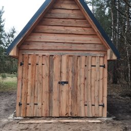 Damian Szlaga - Drewniana szopa z dwuskrzydłowymi drzwiami na tle leśnym, zabezpieczona czarnym zamkiem i zawiasami, z widocznymi śladami piasku wokół podstawy.