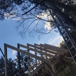 Konstrukcja drewniana dachu w trakcie budowy, widok z dołu na niebieskie niebo z chmurami, z widoczną aluminiową drabiną opartą o drzewo.