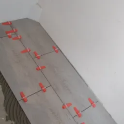 Fragment podłogi w trakcie układania paneli z jasnego drewna, widoczne pomarańczowe kliny dystansowe i pozostałości kleju na podłożu, biała ściana w tle.