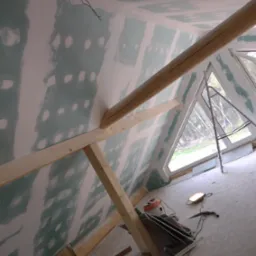 Poddasze w trakcie prac wykończeniowych: widoczne drewniane belki konstrukcyjne, ściany pokryte wstępną warstwą gładzi z widocznymi miejscami po wkrętach, okno trójkątne z widokiem na las, drabina...