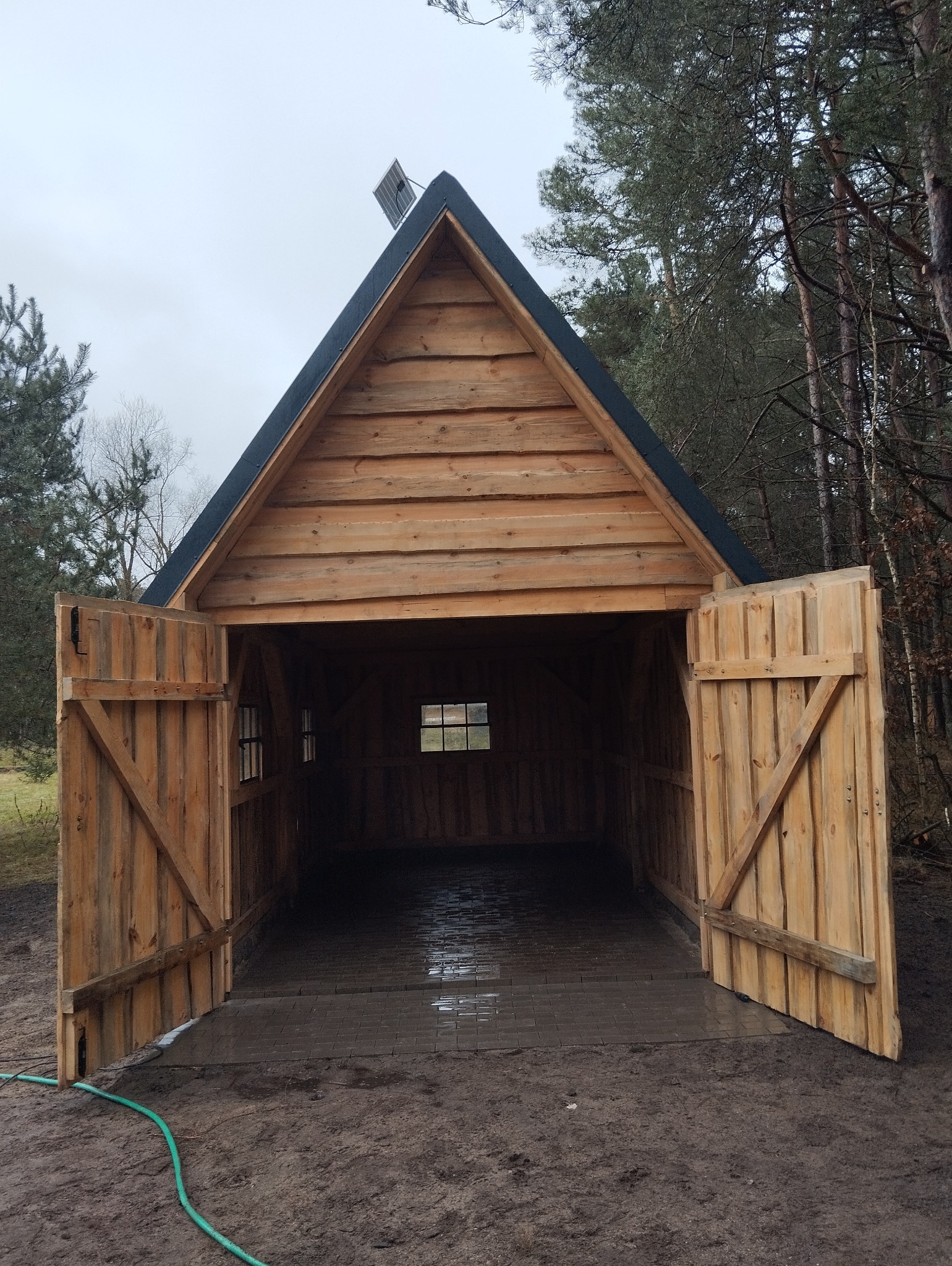 Drewniana wiata z dwuskrzydłowymi drzwiami, wykończona panelami słonecznymi na dachu, na podłożu z kostki brukowej, otoczona lasem.