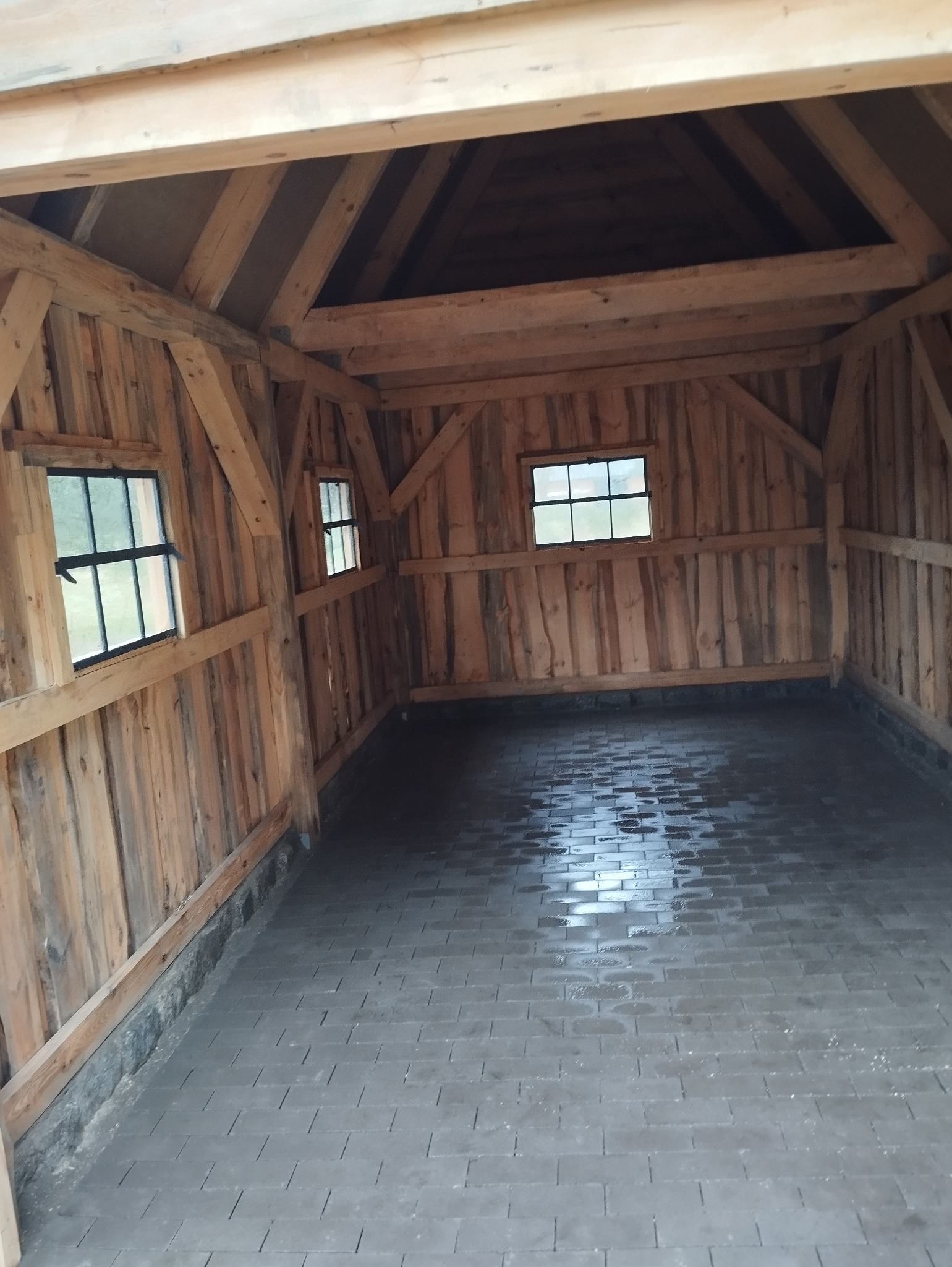 Surowe wnętrze drewnianej konstrukcji z dwoma małymi oknami i posadzką z szarej kostki brukowej, widok z perspektywy wejścia, ukazujący strukturę drewna i minimalizm wykończenia.