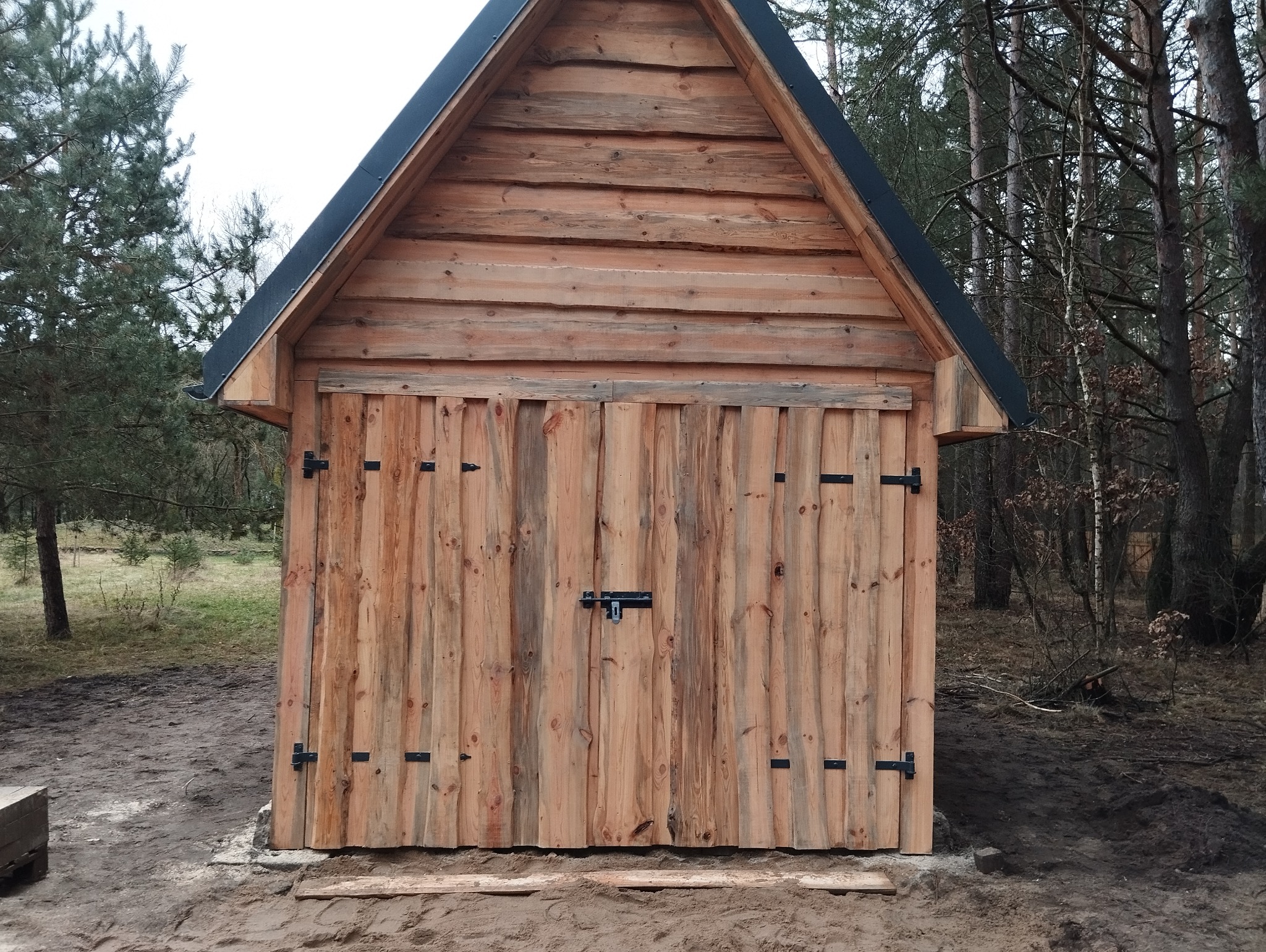 Drewniana szopa z dwuskrzydłowymi drzwiami na tle leśnym, zabezpieczona czarnym zamkiem i zawiasami, z widocznymi śladami piasku wokół podstawy.