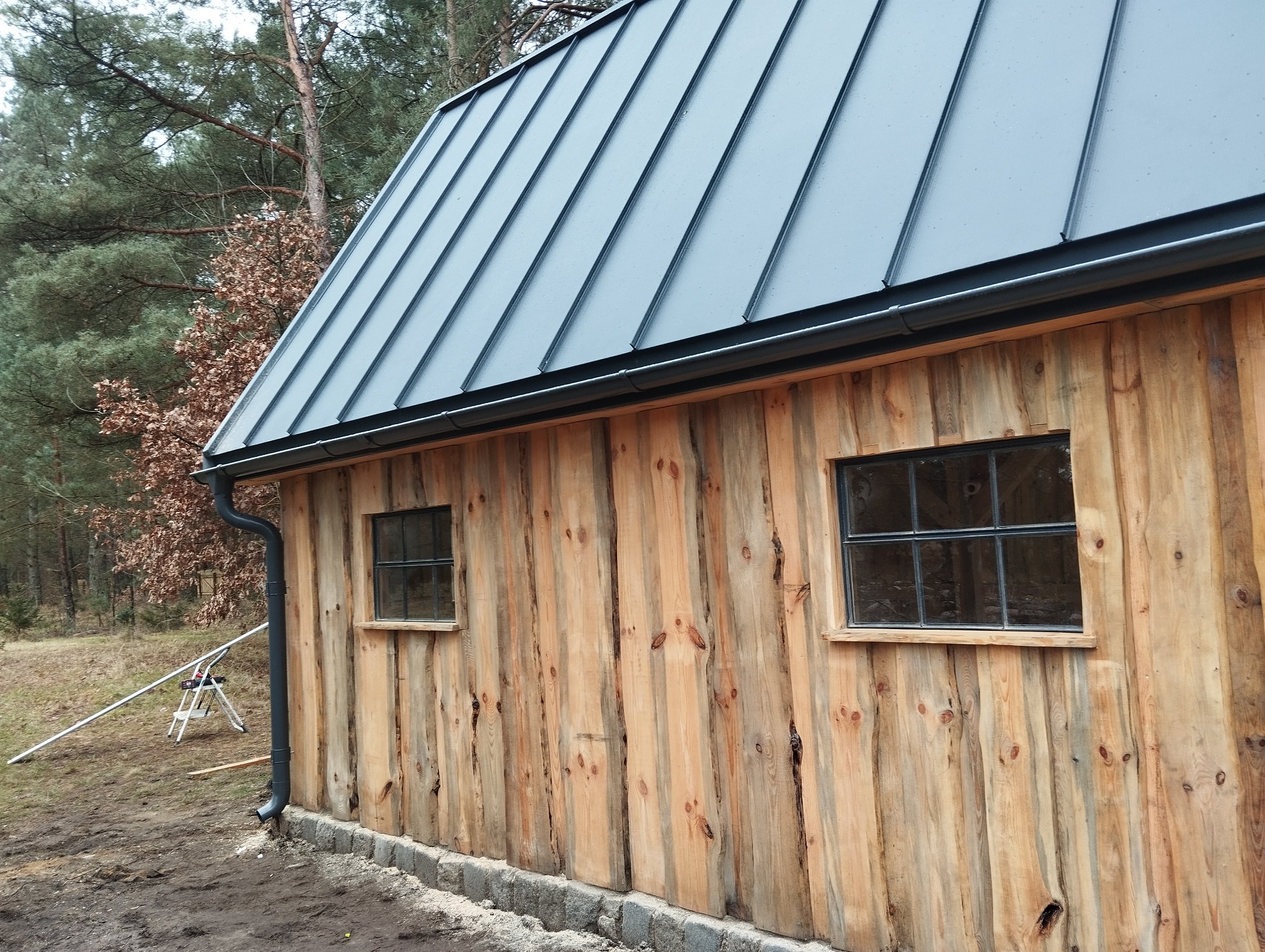Zewnętrzna ściana budynku obita surowym drewnem z widocznymi sękami, szary dach blaszany z orynnowaniem, dwa okna z czarnymi ramami, tło stanowią drzewa.