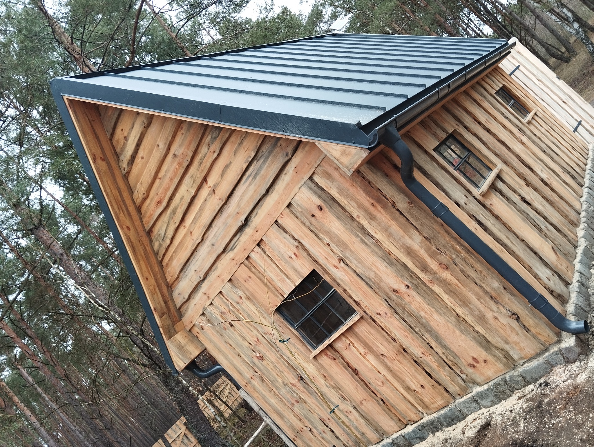 Zewnętrzny widok drewnianego domku z metalowym dachem i czarnymi rynnami, otoczonego drzewami.