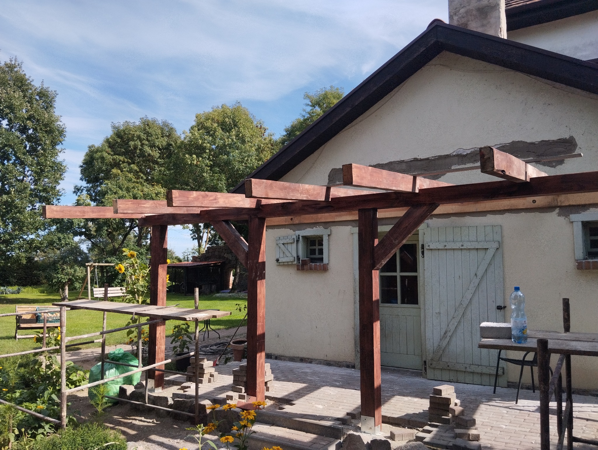 Wznoszona drewniana konstrukcja pergoli przy elewacji domu z widocznymi śladami napraw, narzędzia na prowizorycznym stole, kostka brukowa przygotowana do ułożenia, ogród w tle.