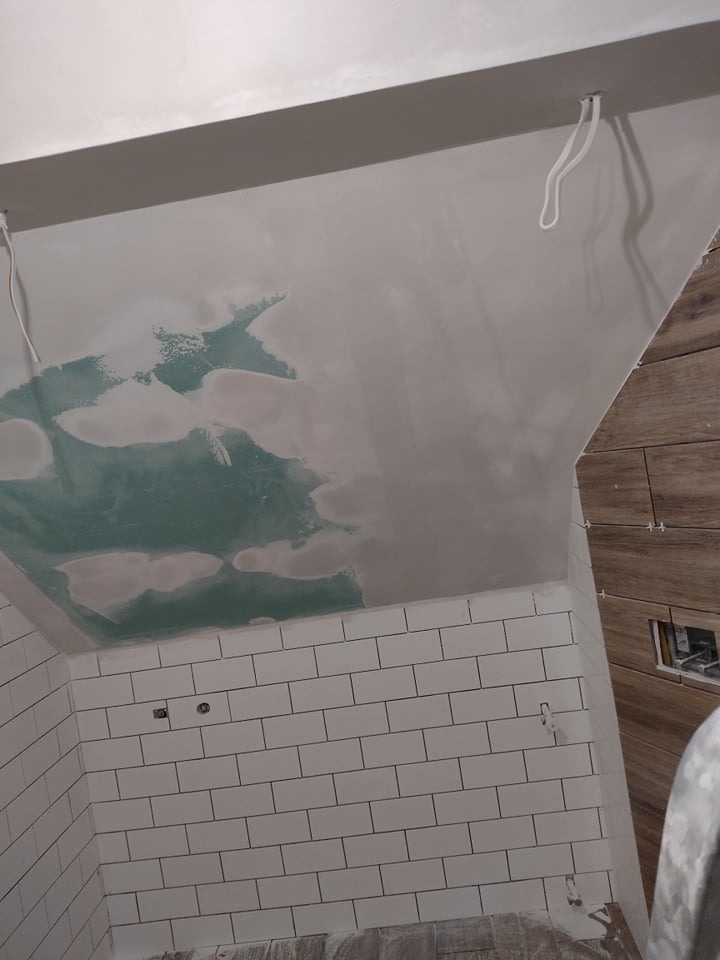 Niedokończone wnętrze łazienki z surowymi, białymi płytkami metra na ścianach i częściowo zagruntowanym sufitem z wystającymi przewodami elektrycznymi, widoczne drewnopodobne płytki na ścianie po...