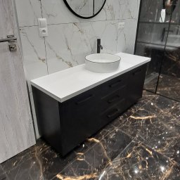 Remigiusz Aksamit - Nowoczesna łazienka z czarną szafką pod umywalkę z białą nablatową umywalką, okrągłym lustrem i marmurowymi płytkami na ścianie i podłodze.