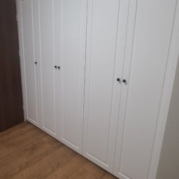 Remigiusz Aksamit - Biała szafa wnękowa z frezowanymi frontami i okrągłymi uchwytami, dopasowana do sufitu, na podłodze panele w odcieniach brązu. Widok z dołu, drzwi zamknięte.