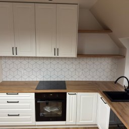 Remigiusz Aksamit - Biała zabudowa kuchenna na poddaszu z drewnianym blatem i geometrycznym wzorem na ścianie. Widoczna czarna armatura i płyta indukcyjna.