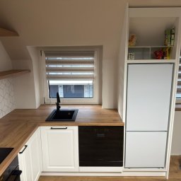 Remigiusz Aksamit - Funkcjonalna kuchnia na poddaszu w Opole. Białe szafki z drewnianym blatem, czarny zlew i AGD. Zabudowa lodówki z półkami, okno z żaluzjami.