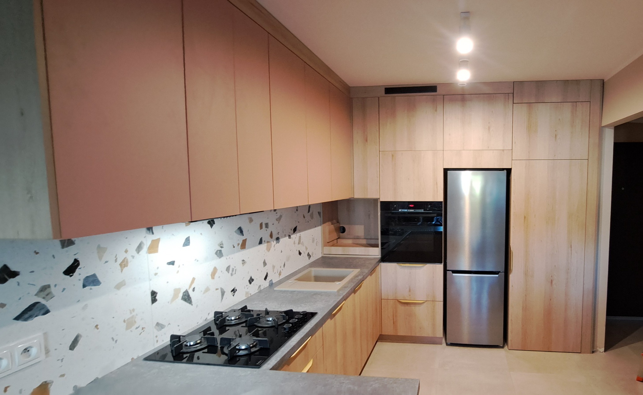 Jasna kuchnia z drewnianymi frontami, blatem w odcieniach szarości i mozaiką na ścianie, z widoczną płytą gazową i lodówką.