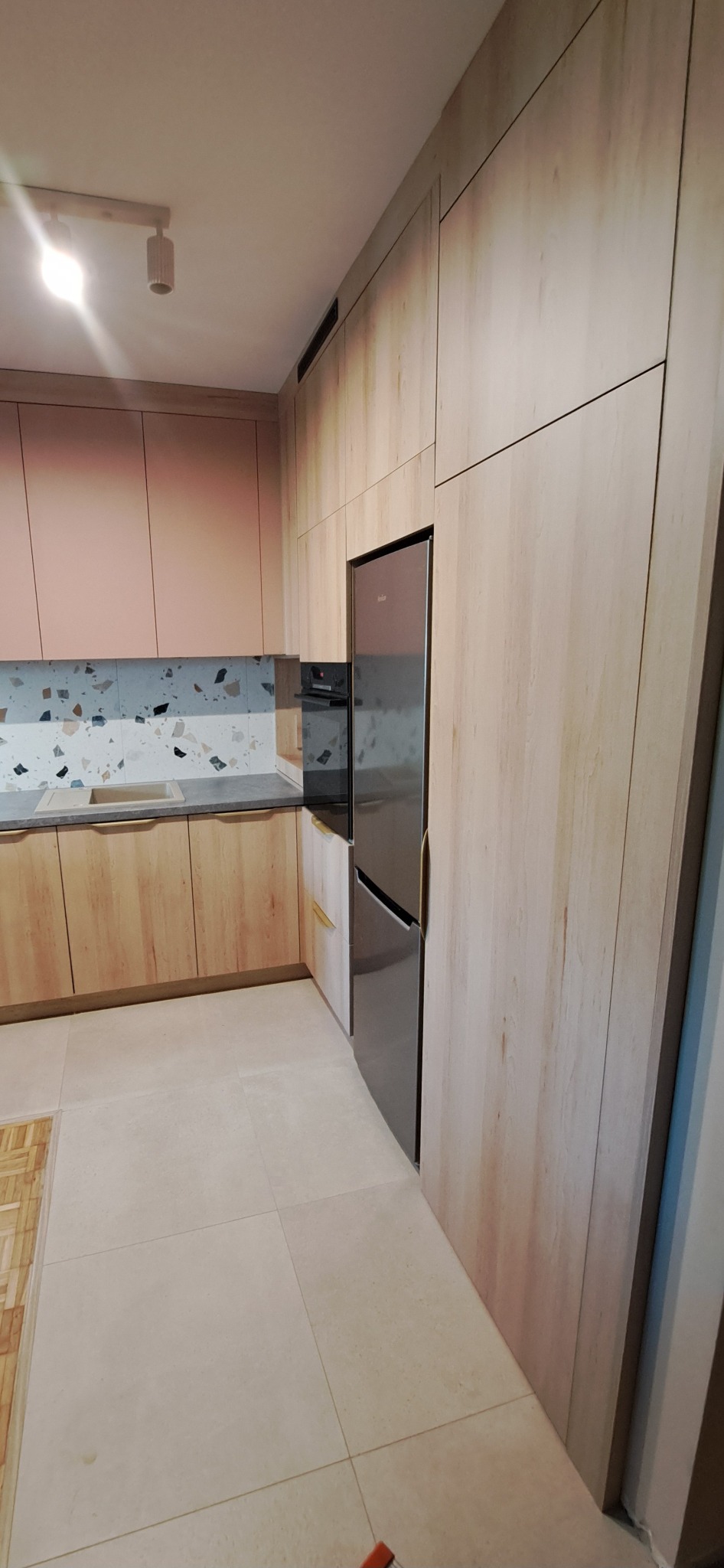 Jasna kuchnia z drewnianymi frontami, różowymi szafkami górnymi i mozaiką nad blatem, z wbudowaną lodówką i piekarnikiem.