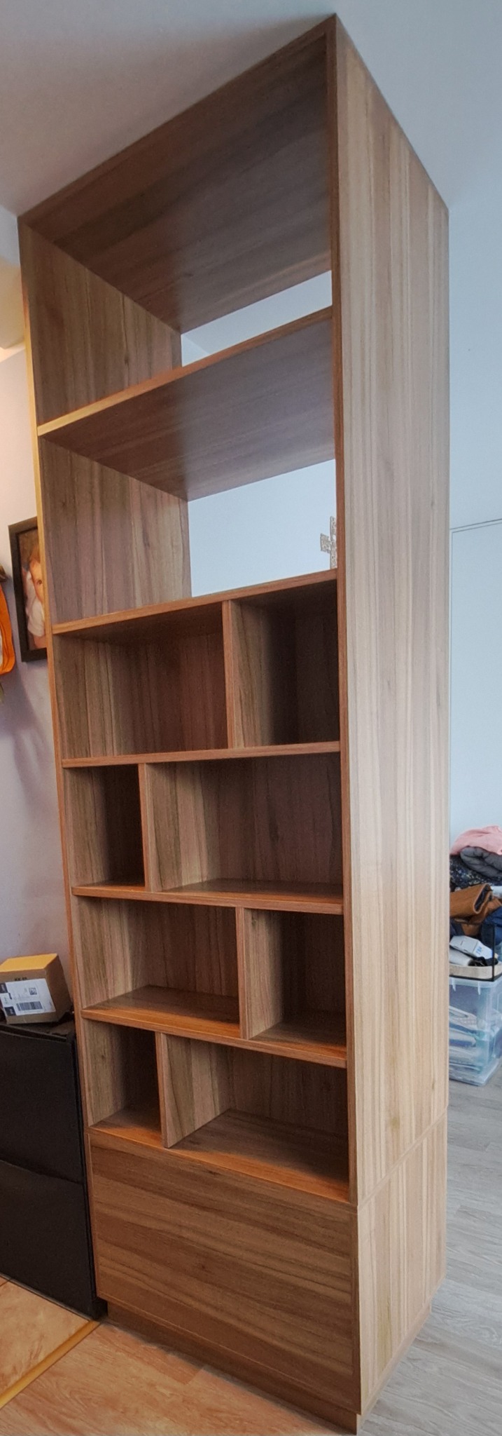 Wysoka, drewniana szafa na wymiar z otwartymi półkami i szufladą na dole, wykonana w jasnym odcieniu drewna, stojąca w pokoju.