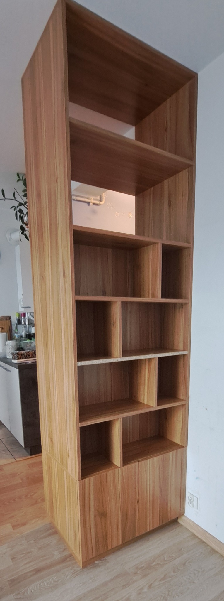 Wysoka, drewniana szafa z półkami i szafką na dole, z widoczną strukturą drewna, ustawiona w jasnym wnętrzu. Widoczne detale wykończenia.