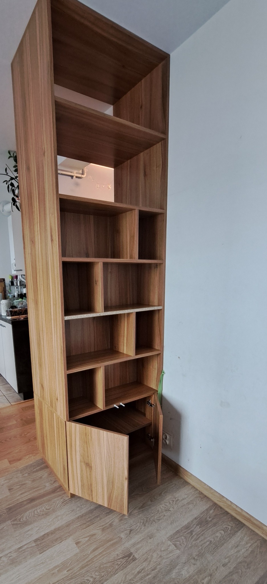 Wysoka, drewniana szafa z otwartymi półkami i dolną szafką z uchylonymi drzwiczkami, dopasowana do rogu pokoju z widoczną instalacją elektryczną.