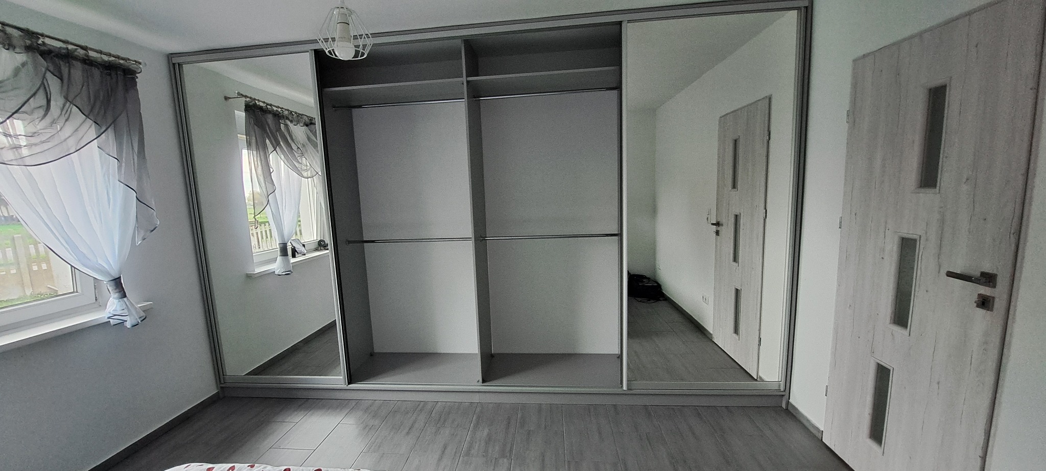 Jasna szafa wnękowa z przesuwnymi drzwiami lustrzanymi, widok na wnętrze z półkami i drążkami, szare panele tylne. Widok na okno i drzwi w odbiciu.