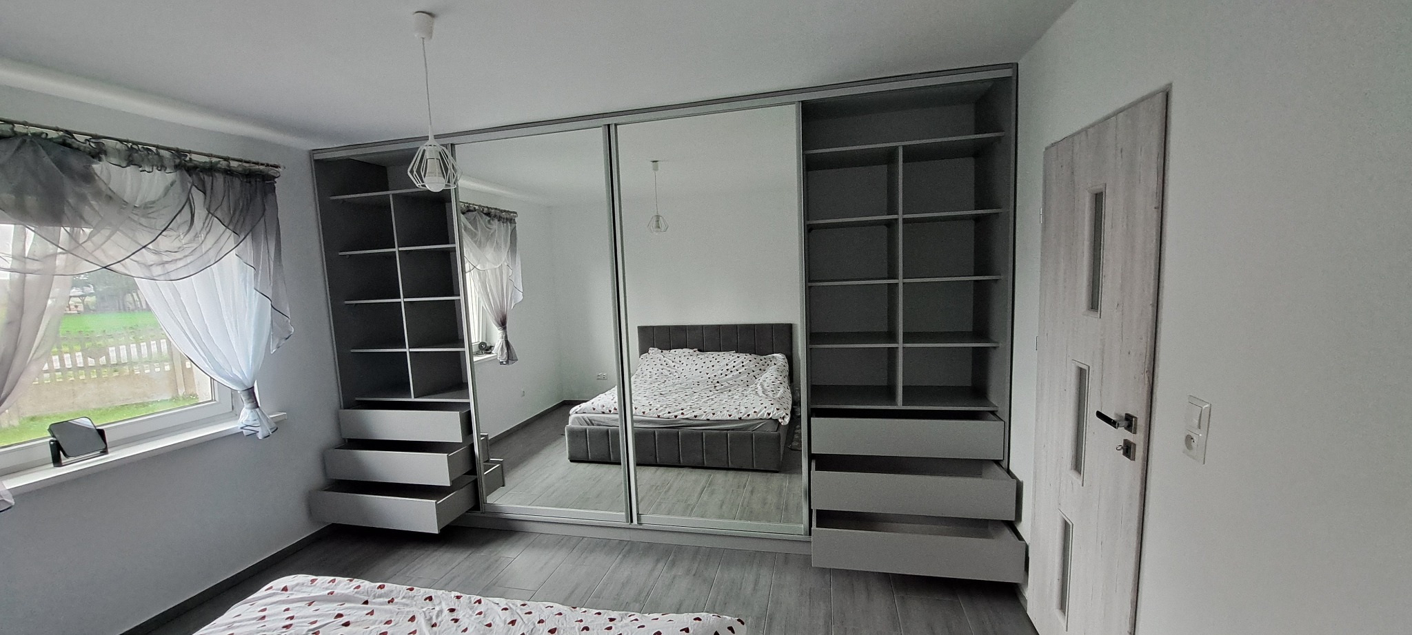 Szara szafa wnękowa z lustrzanymi drzwiami przesuwnymi w sypialni, z otwartymi szufladami i półkami po bokach. Widoczne odbicie łóżka i okna.