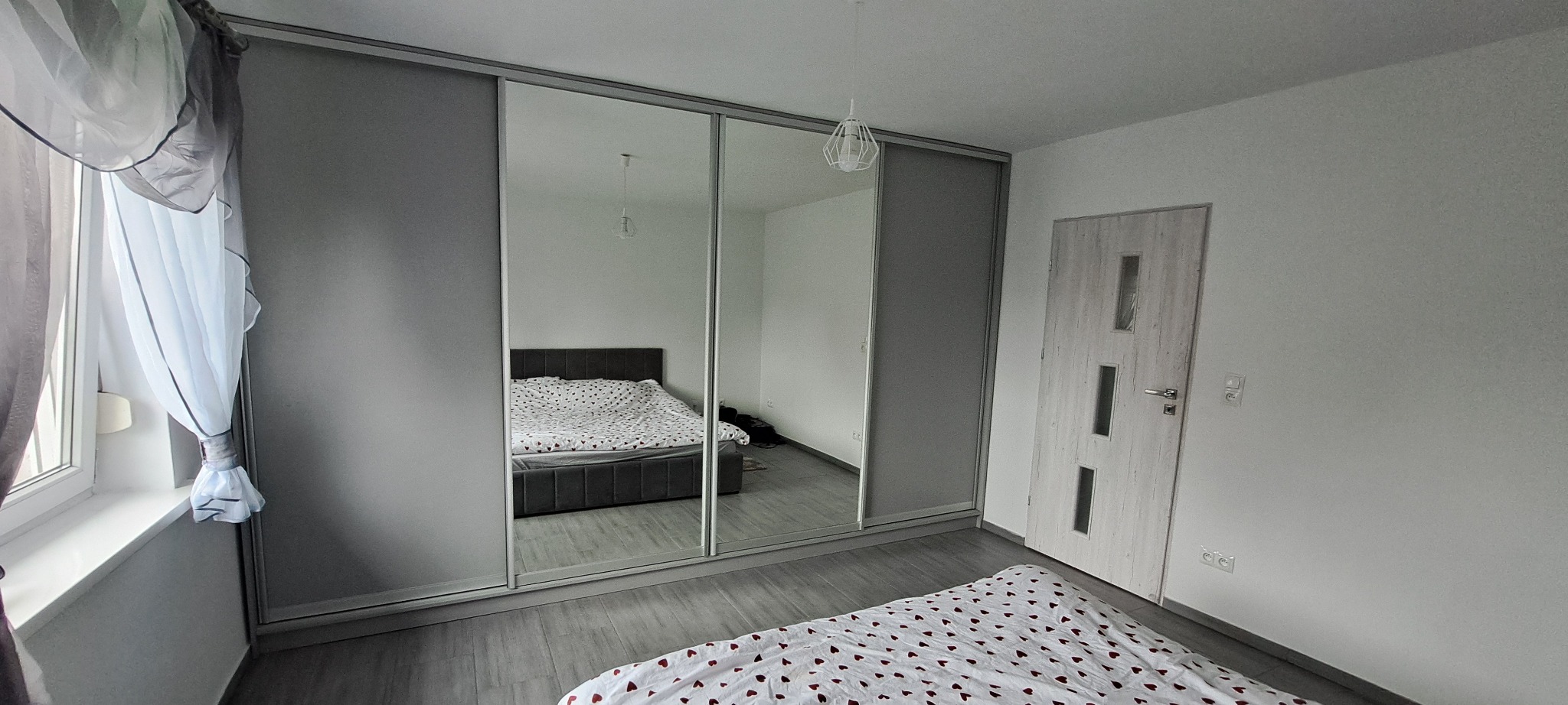 Nowoczesna szafa wnękowa z przesuwnymi, lustrzanymi drzwiami w sypialni z łóżkiem odbitym w lustrze. Szare panele boczne i białe ściany.