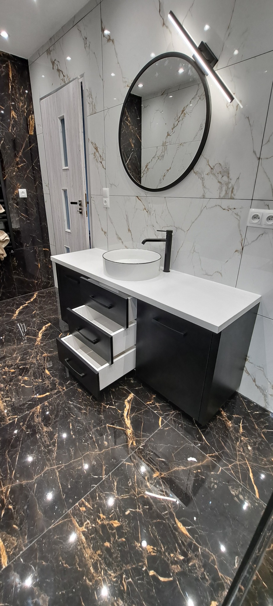 Nowoczesna szafka łazienkowa na wymiar w czerni i bieli, z okrągłym lustrem i otwartymi szufladami, na tle marmurowych płytek. Eleganckie wykończenie.