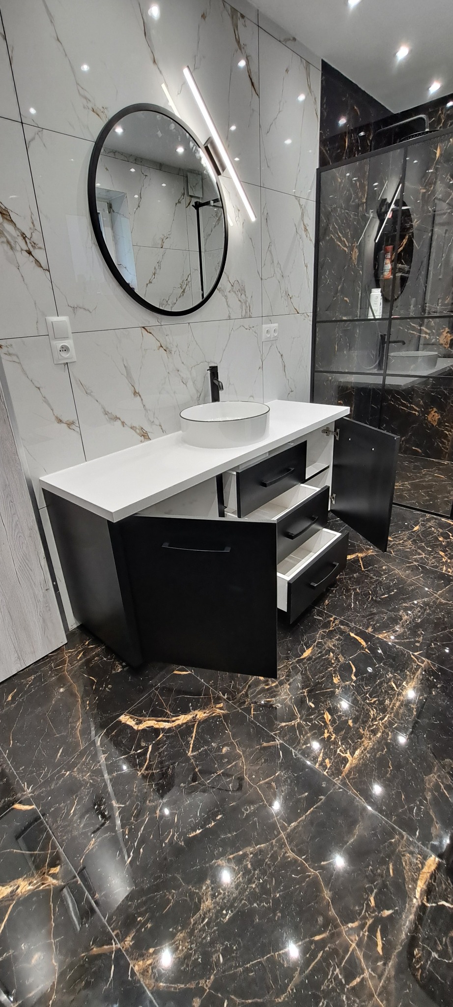 Czarna szafka łazienkowa z białym blatem i okrągłym lustrem na ścianie z marmurowym wzorem. Otwarta szafka ukazuje szuflady. Podłoga z ciemnego marmuru.