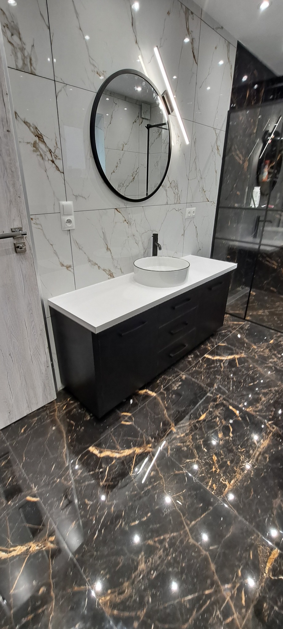 Nowoczesna łazienka z czarną szafką pod umywalkę z białą nablatową umywalką, okrągłym lustrem i marmurowymi płytkami na ścianie i podłodze.