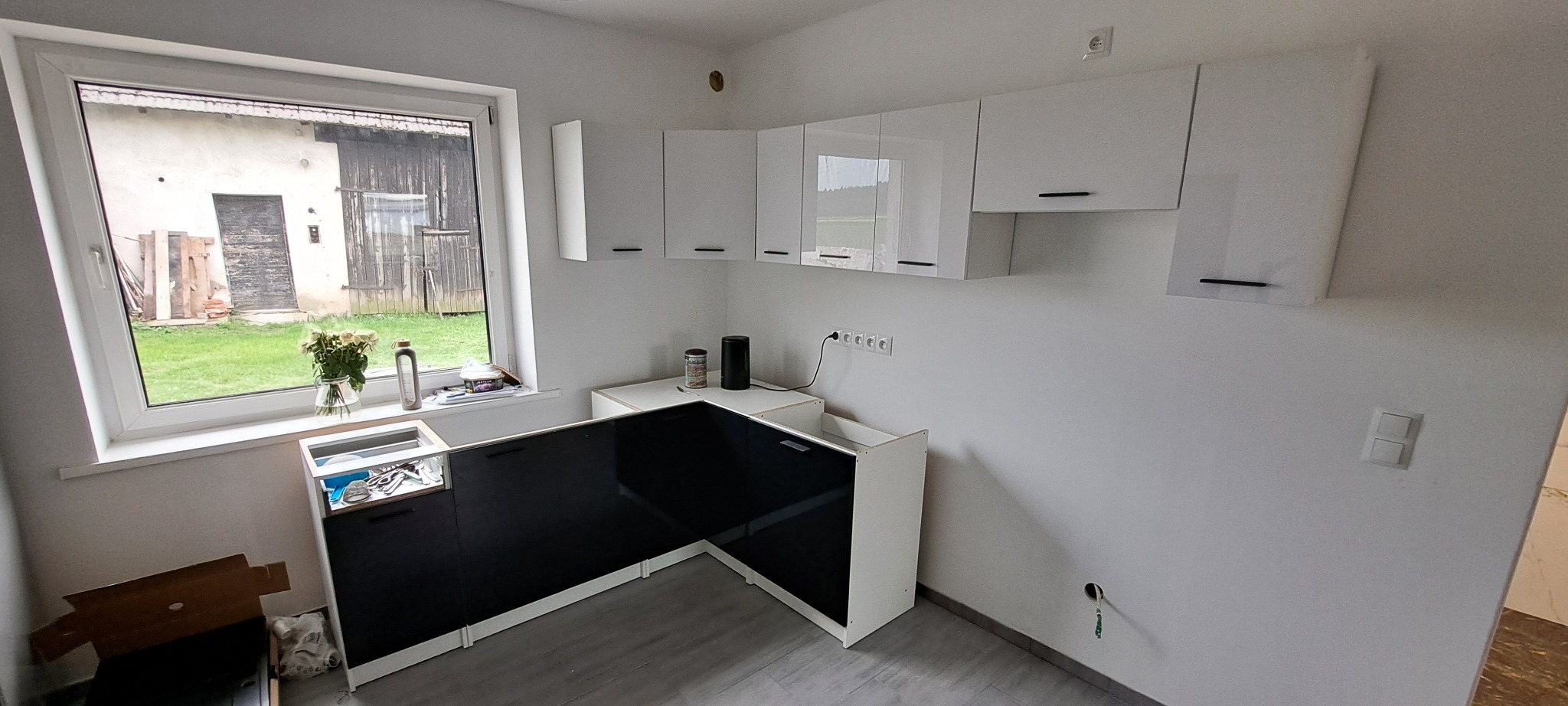 Częściowo zamontowana, nowoczesna kuchnia z czarnymi frontami i białymi szafkami wiszącymi. Widok na zewnątrz przez okno.