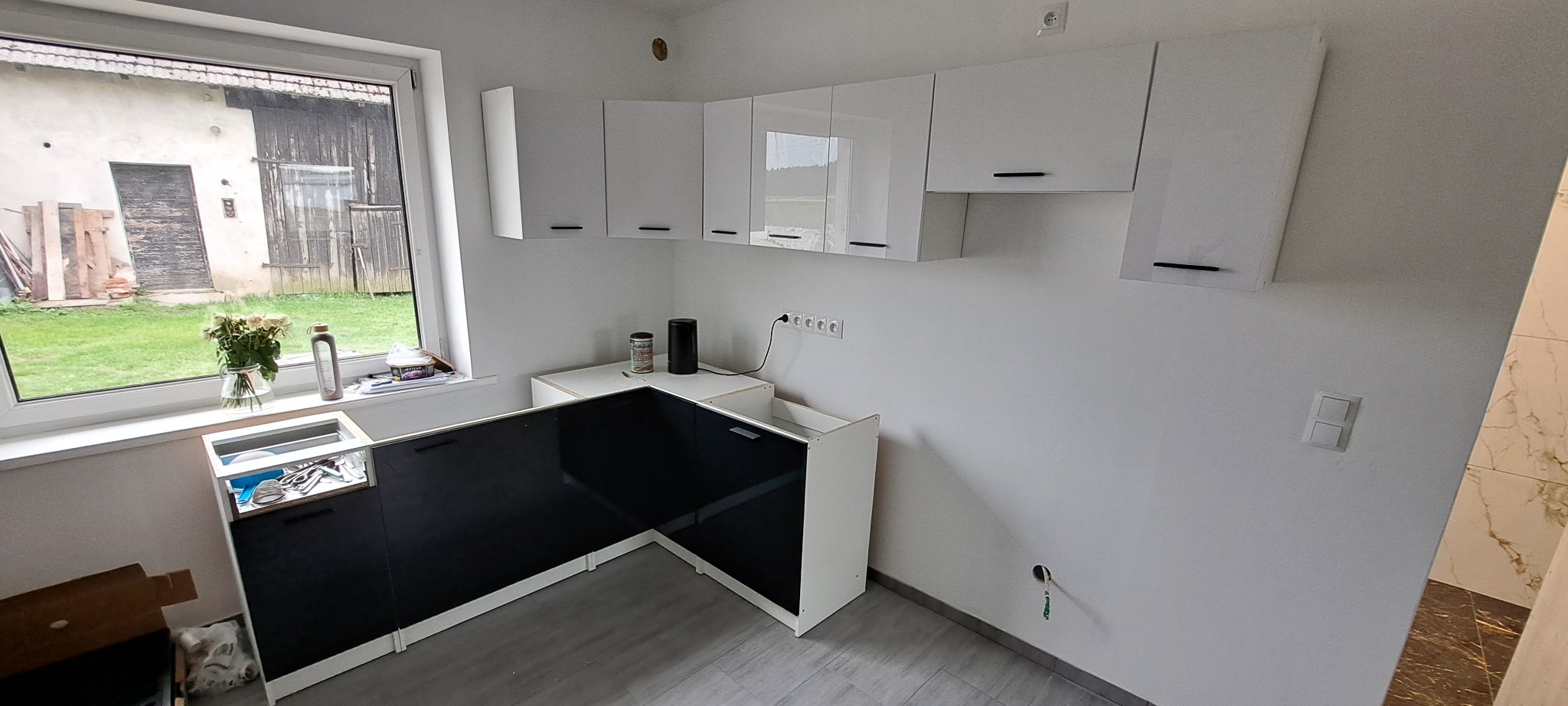 Częściowo zmontowana kuchnia z czarnymi frontami i białymi szafkami wiszącymi. Widok na wiejską zabudowę przez okno.