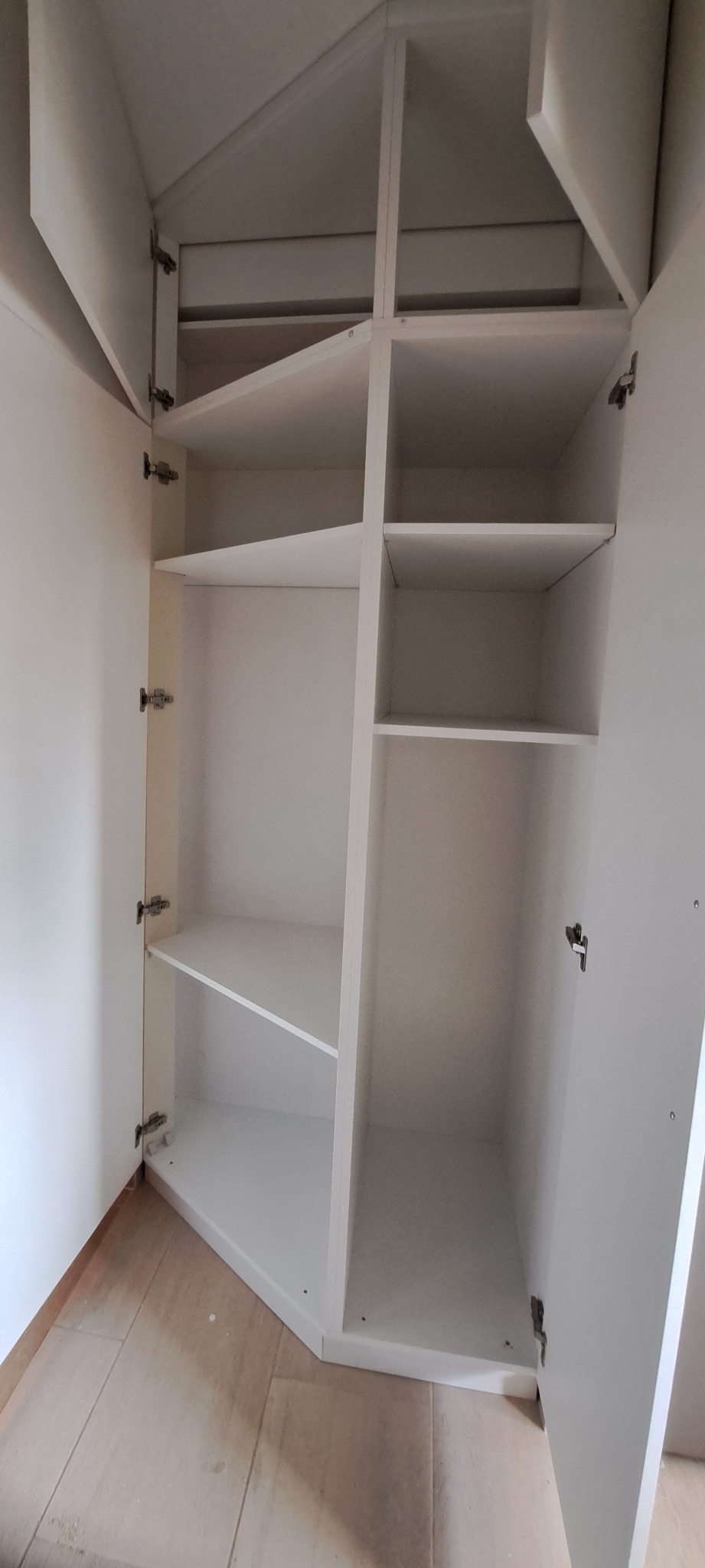 Wnętrze białej szafy wnękowej z półkami, widok z dołu, drzwi otwarte, widoczne zawiasy, jasne drewniane panele na podłodze, oświetlenie dzienne.