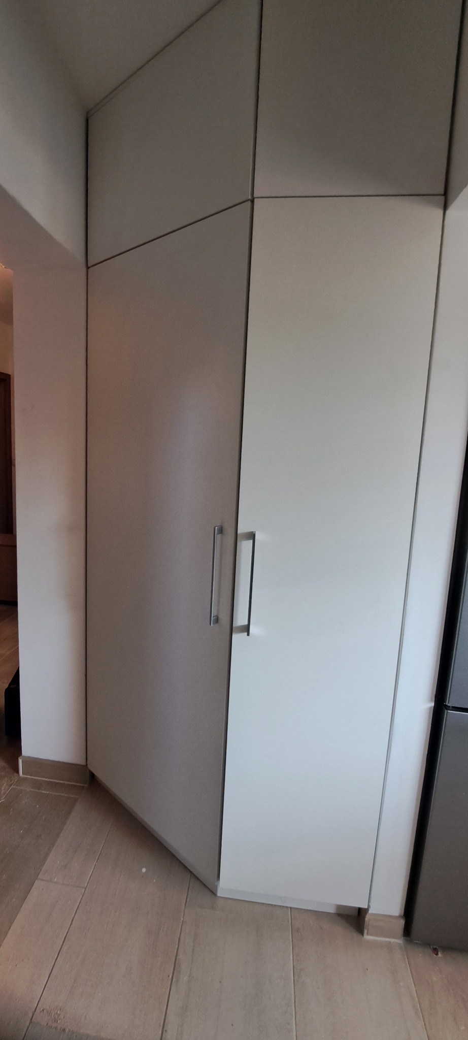 Szara szafa wnękowa z białymi drzwiami i długimi, prostymi uchwytami, dopasowana do rogu pomieszczenia z drewnianą podłogą.