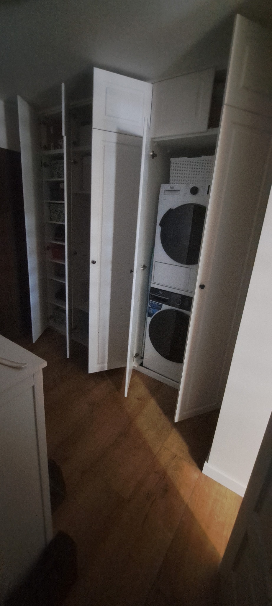 Biała szafa wnękowa z otwartymi drzwiami, ukazująca półki i pralko-suszarkę. Widoczne drewniane panele podłogowe i fragment białej komody.