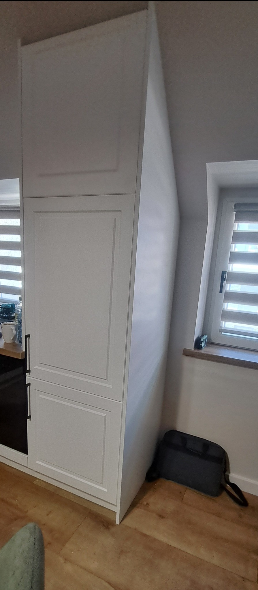 Biała szafa wnękowa z frezowanymi frontami, dopasowana do skosu poddasza, obok okna z żaluzjami i torba narzędziowa na podłodze.