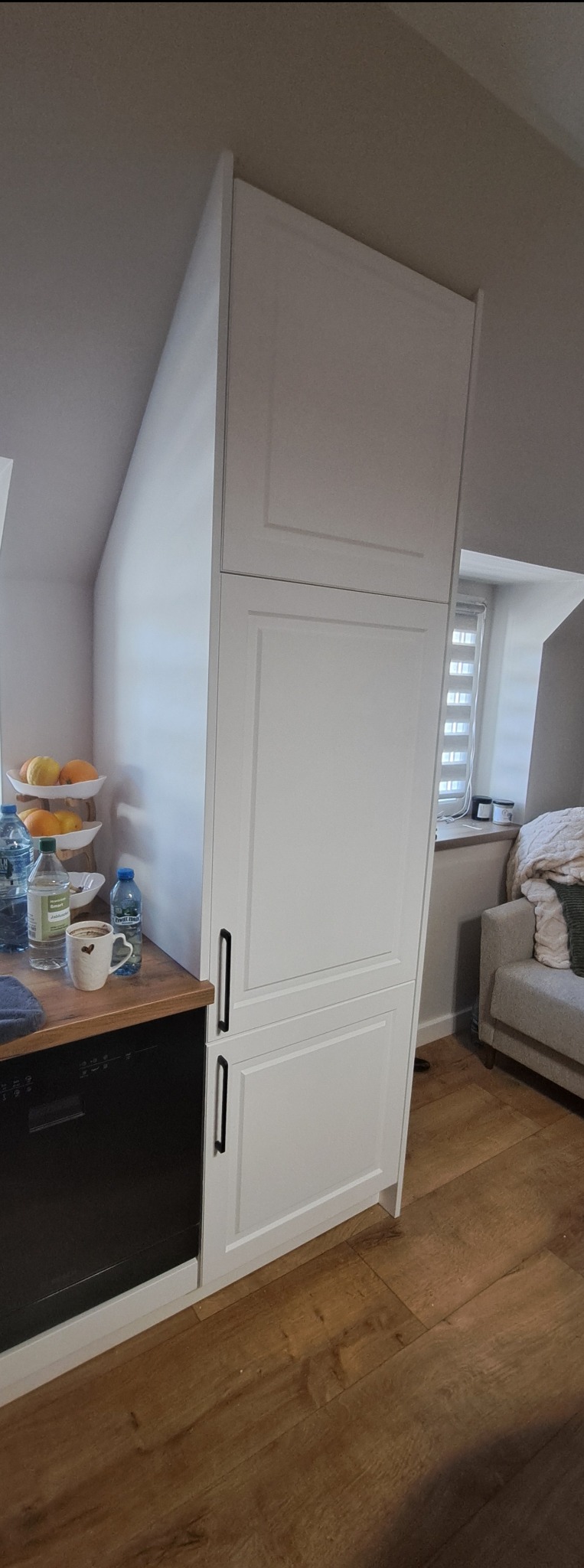 Biała szafa kuchenna na wymiar z czarnymi uchwytami, dopasowana do skosu poddasza. Obok blat z owocami i napojami, w tle okno i sofa.