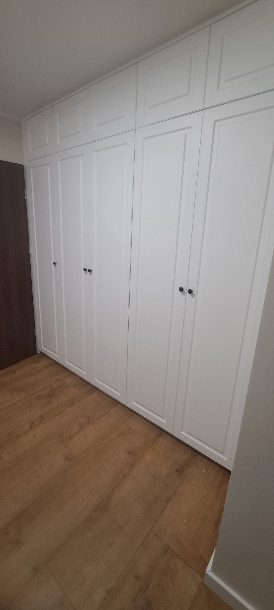 Biała szafa wnękowa z frezowanymi frontami i okrągłymi uchwytami, dopasowana do sufitu, na podłodze panele w odcieniach brązu. Widok z dołu, drzwi zamknięte.