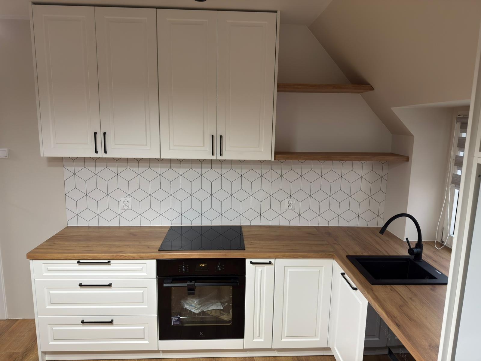 Biała zabudowa kuchenna na poddaszu z drewnianym blatem i geometrycznym wzorem na ścianie. Widoczna czarna armatura i płyta indukcyjna.