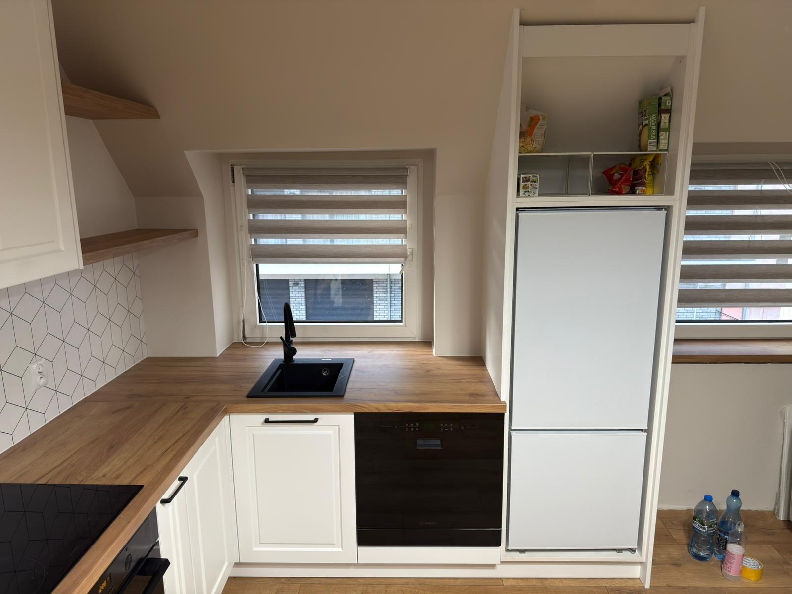 Funkcjonalna kuchnia na poddaszu w Opole. Białe szafki z drewnianym blatem, czarny zlew i AGD. Zabudowa lodówki z półkami, okno z żaluzjami.