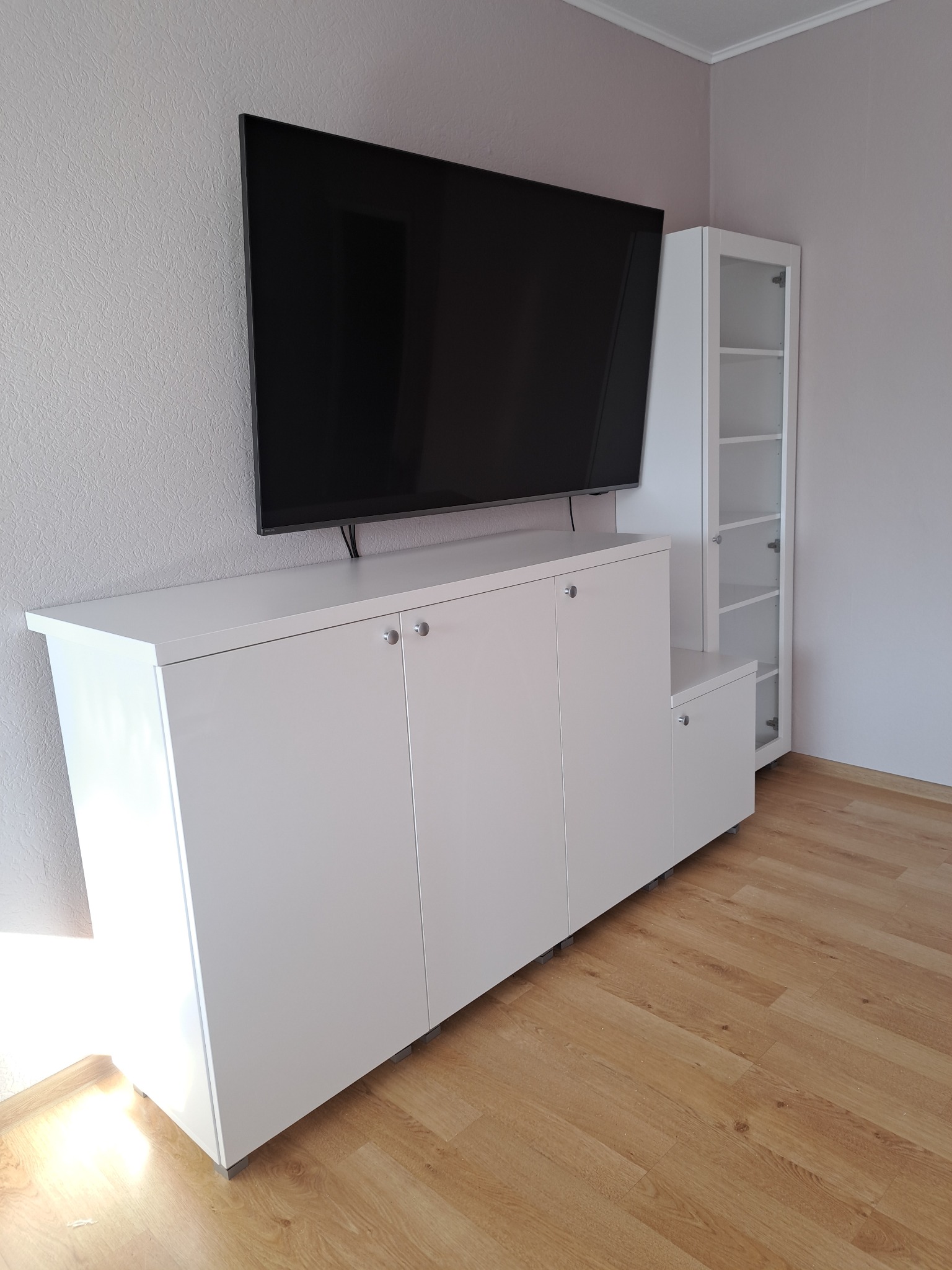 Biała szafka RTV pod telewizorem, obok regał z półkami, na drewnianej podłodze. Minimalistyczny design, jasne wnętrze, nowoczesny styl.