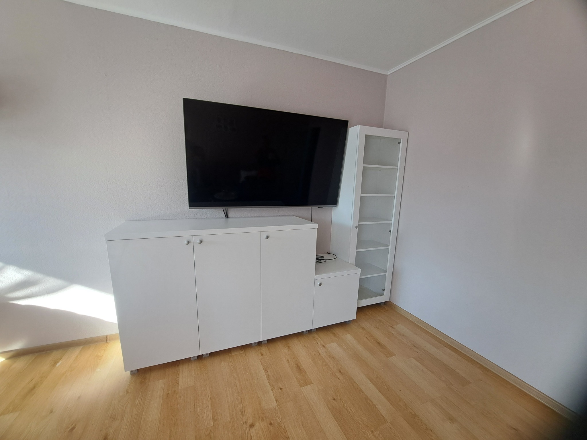 Biała szafka RTV z szafką i półką, ustawiona pod telewizorem na ścianie w pokoju o jasnych ścianach i parkiecie. Minimalistyczny design.