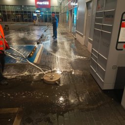 Z POŁYSKIEM Bartłomiej Zienkiewicz - Dwóch mężczyzn czyści kostkę brukową przed budynkiem sklepu Martex i automatem DPD. Jeden używa specjalnej maszyny, drugi lancy ciśnieniowej, widać mokrą powierzchnię.